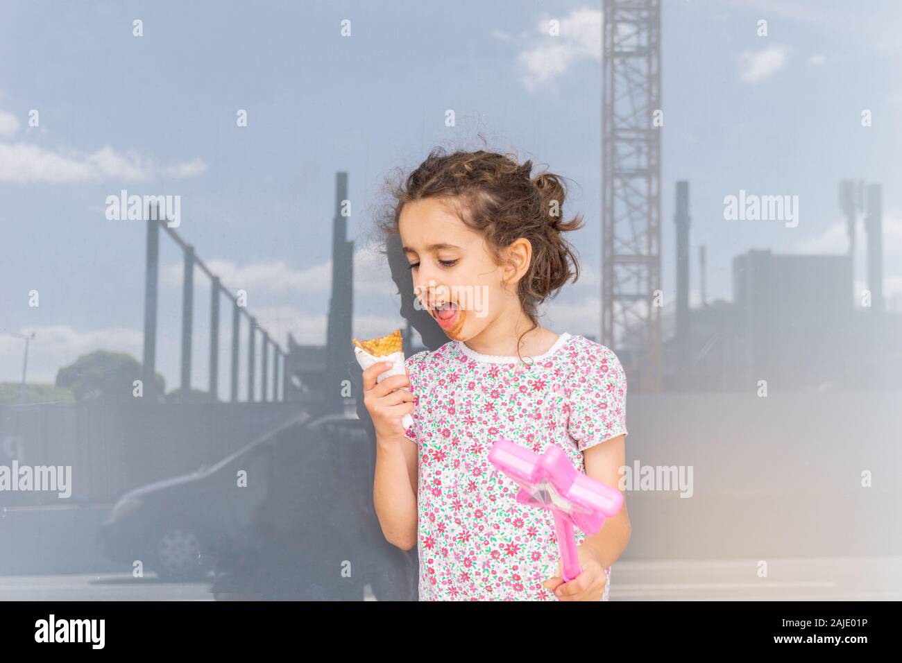 Kleines Mädchen mit unordentlichen Gesicht genießen Sie Schokolade essen halten rosa Zauberstab mit Stern in der Stadt mit dem Bau Skyline spiegelt sich in Fenster hinter. Stockfoto