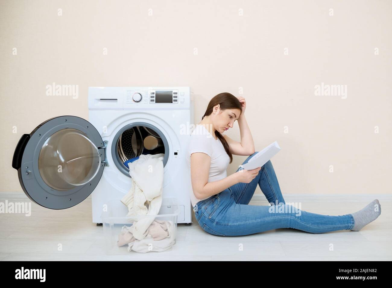 Junge Hausfrau lernt Einstellungen der neuen Waschmaschine mit Anweisung sitzen auf dem Boden im Zimmer Stockfoto