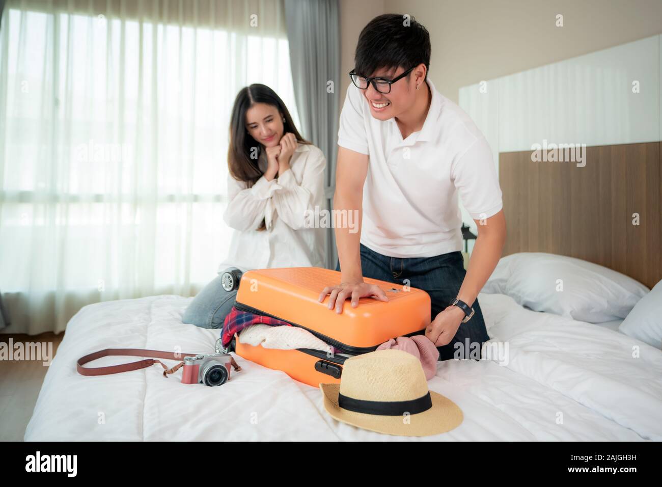 Asiatischer Mann, der versucht, alle Kleidung zu verpacken seiner orange Koffer vor dem Urlaub und ihre Freundin auf dem Bett im Hintergrund passt. im Schlafzimmer an Stockfoto Asiatischer Mann, der versucht, alle Kleidung zu verpacken seiner orange Koffer vor dem Urlaub und ihre Freundin auf dem Bett im Hintergrund passt. im Schlafzimmer an Stockfoto