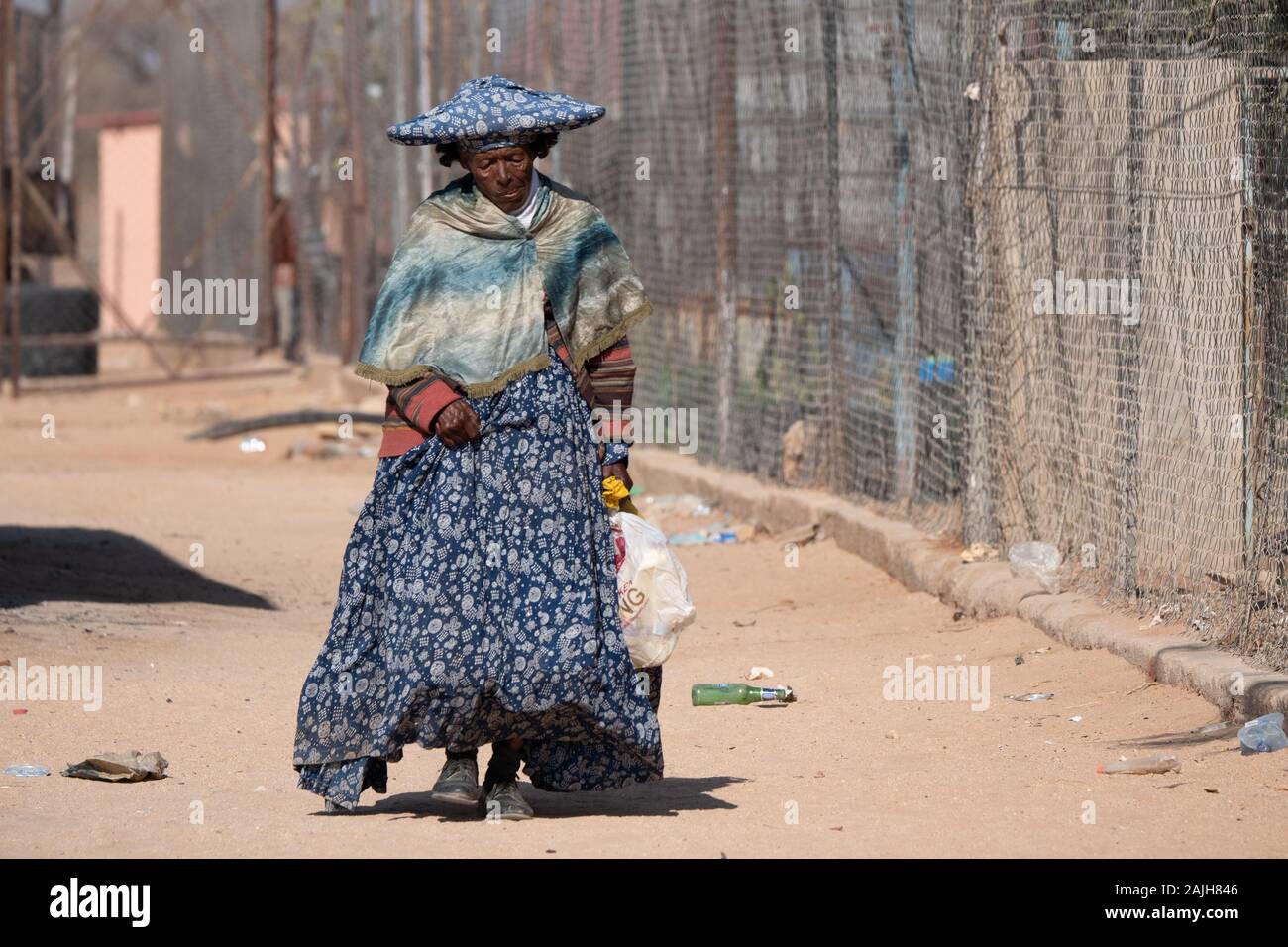 Omatjette, Erongo Region, Namibia - 24. Juli 2019: armen alten Herero Frau in traditioneller Kleidung mit Horn-Shaped hat man durch eine verschmutzte Neighborhoo Stockfoto