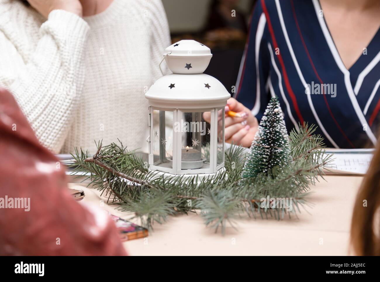 Ein weißes Metall Leuchter mit Kerze innen mit Weihnachtsbaum umgeben. Frauen sitzen um den Tisch. Weihnachtsfeier oder treffen. Stockfoto