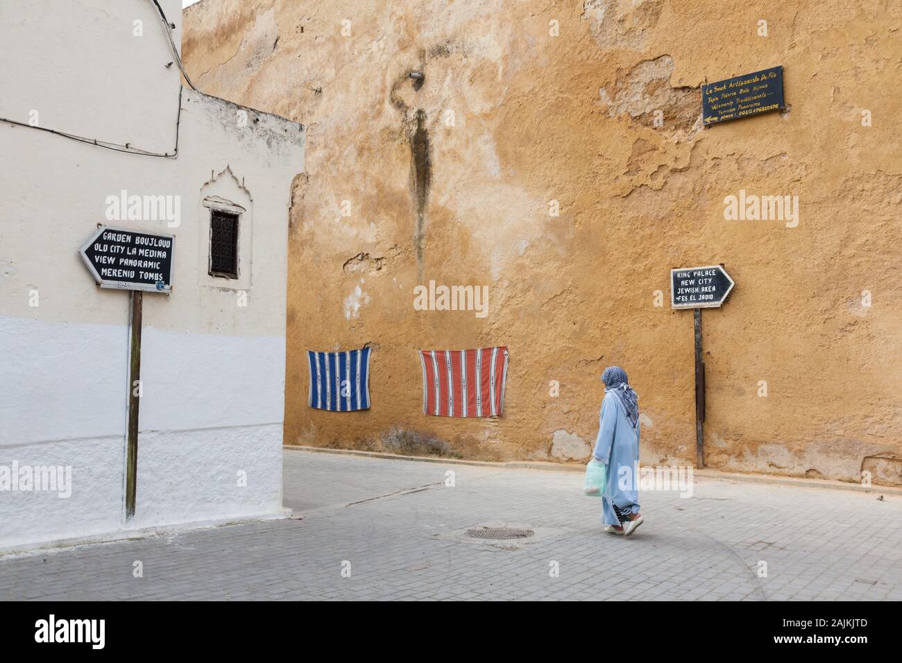 Frau in blauem Kleid und mit Kopftuch am Kopf, Wegweiser zu Touristenattraktionen und Stoffen, die an der Wand hängen, Fes (Fez), Marokko Stockfoto