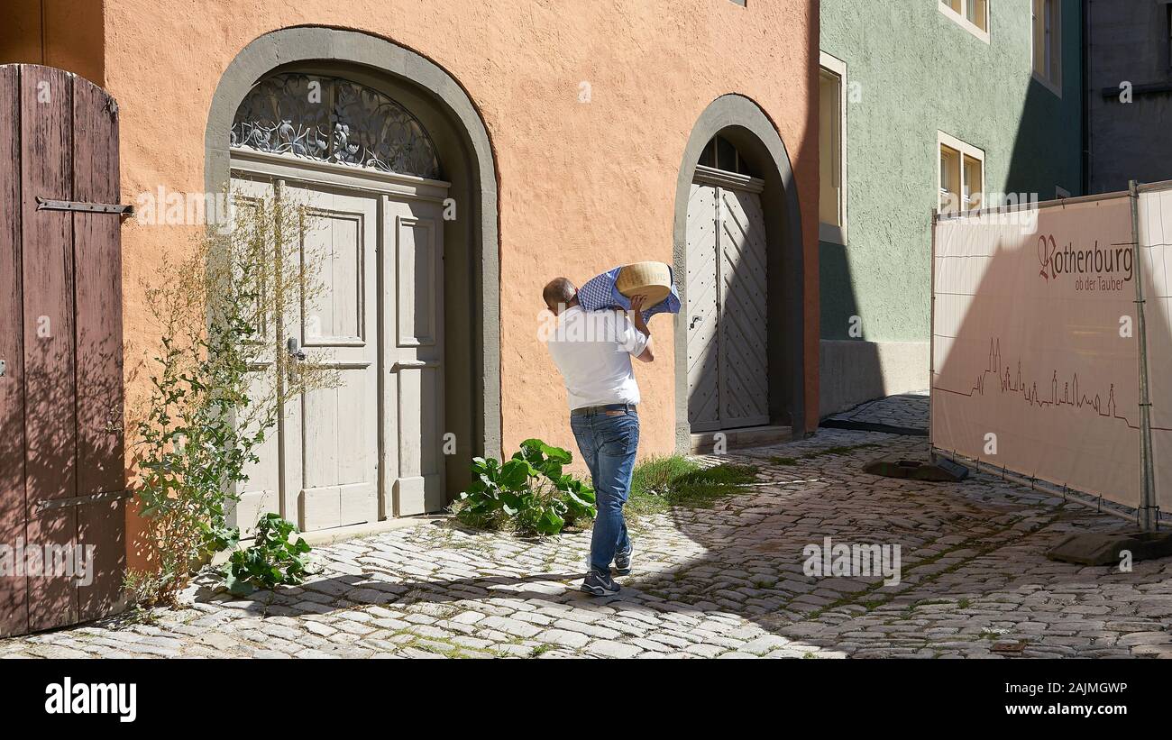 Der Mensch trägt große gourmet Käse Rad auf seine Schulter, als er die Auslieferung zu einem Shop in einer schmalen Straße mit Kopfsteinpflaster im mittelalterlichen Rothenburg macht, Deutschland Stockfoto