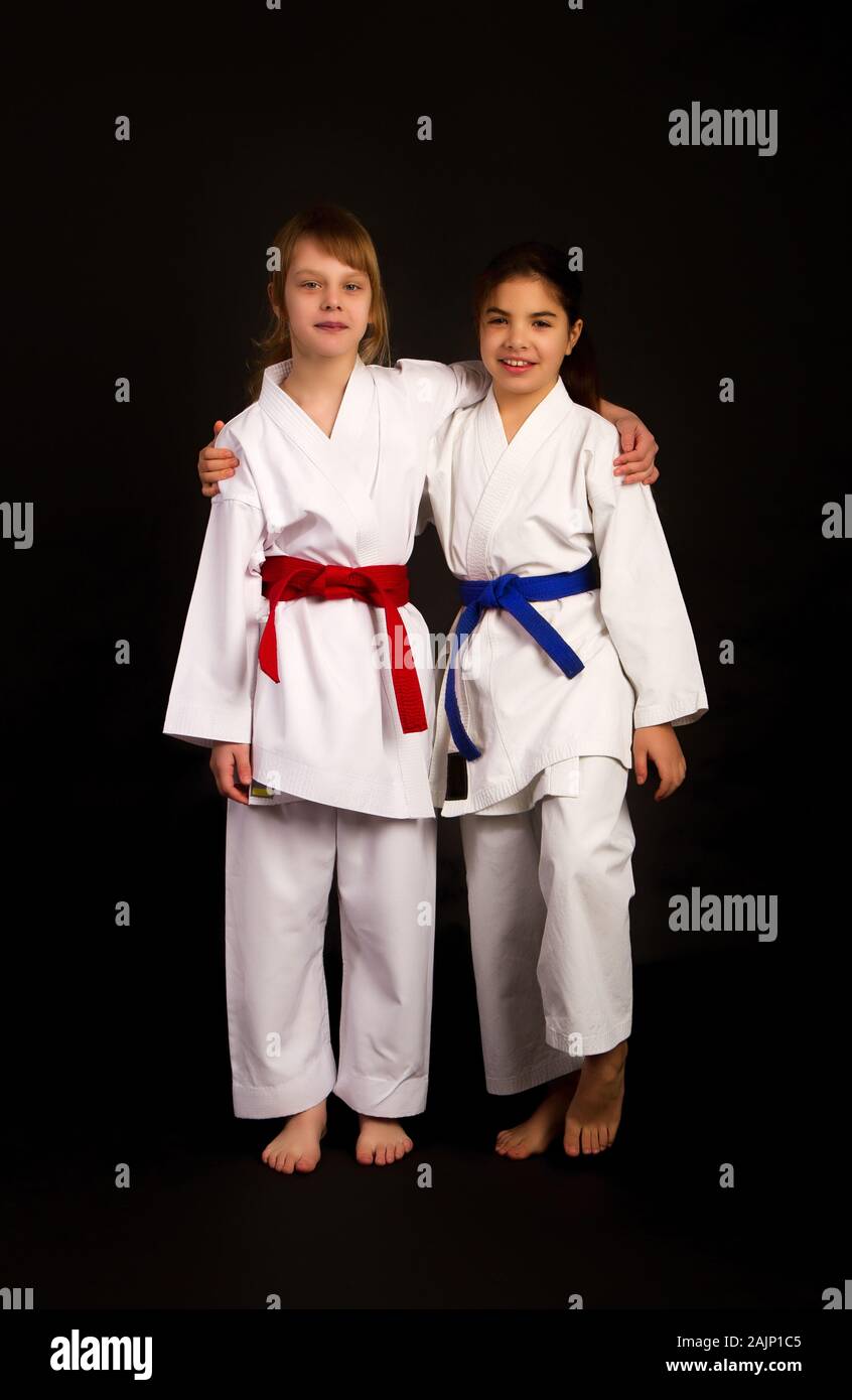 Zwei kleine Karate Mädchen Freunde in Weiß Kimonos, eine in Rot und die andere blau Wettbewerb Riemen umarmen vor einem dunklen Hintergrund Stockfoto