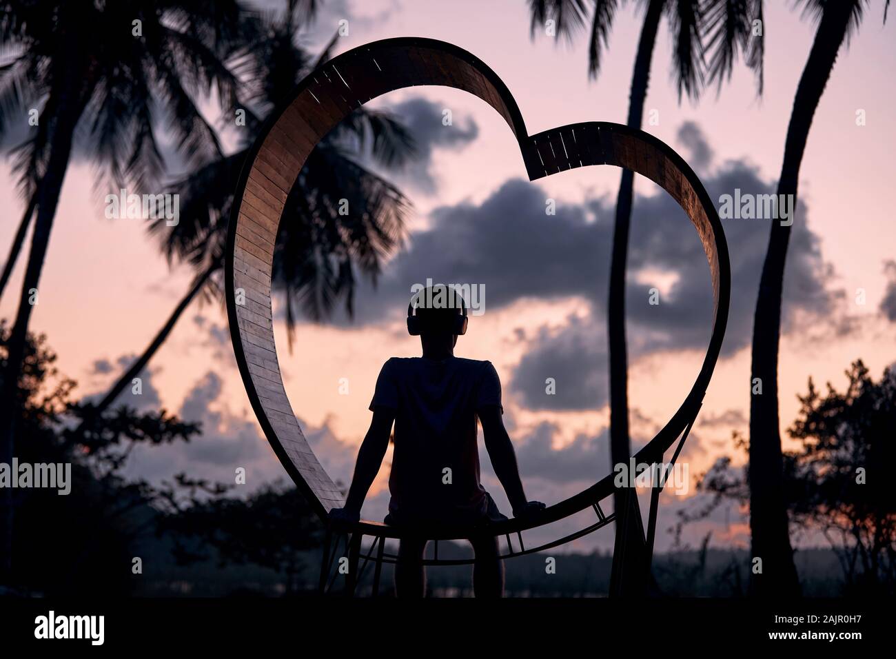 Entspannen im tropischen Paradies. Junger Mann mit Kopfhörern in Holz- Herzen sitzen und hören Musik gegen die Palmen bei Sonnenuntergang. Stockfoto