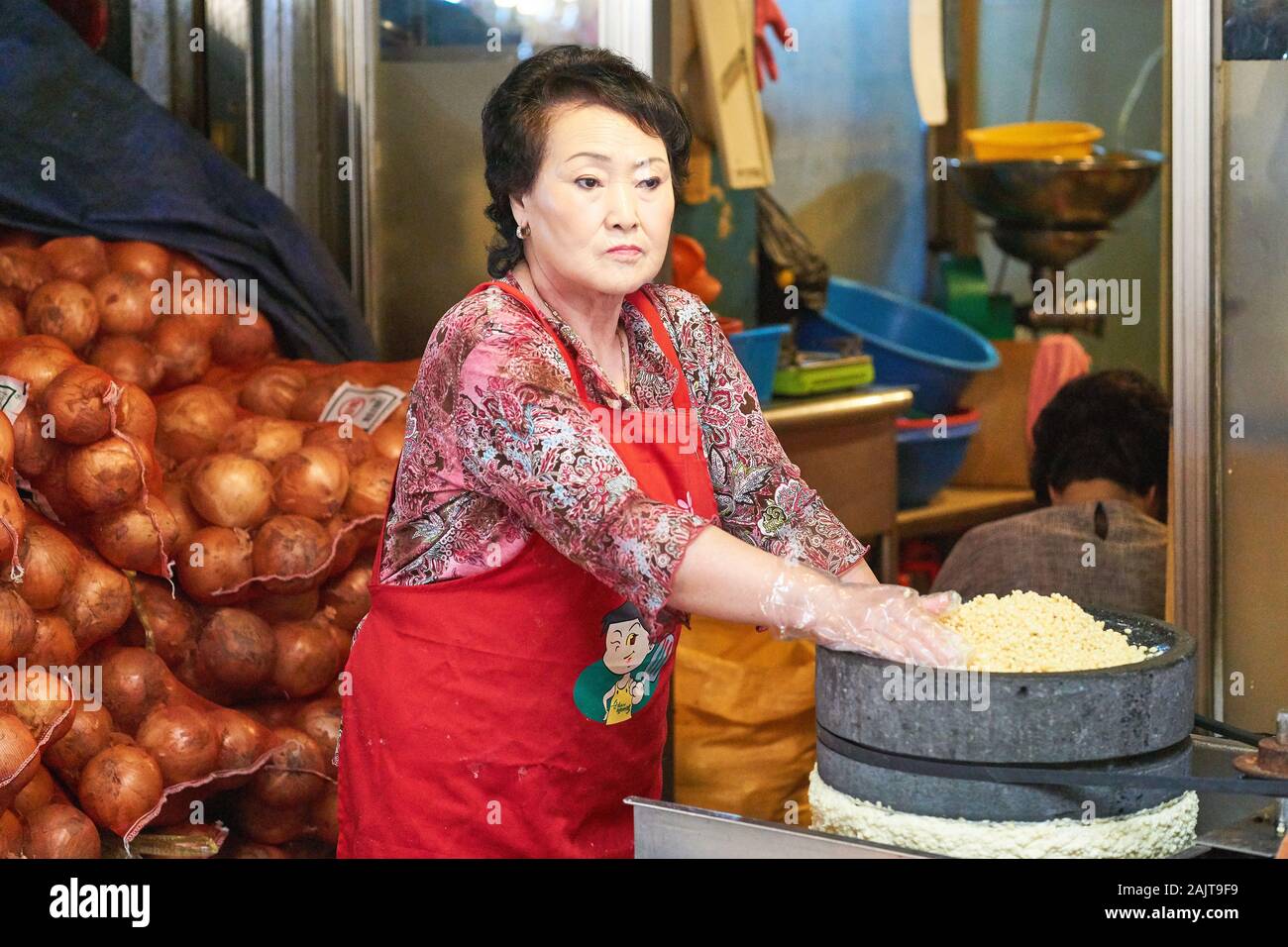 Eine Koreanerin in roter Schürze verwendet eine spezielle Mühle, um Mungbohnen für die Verwendung in Mungbohnenpfannkuchen (Bindae-tteok oder Bindaetteok) im Gwangjang MKT zuzubereiten. Stockfoto