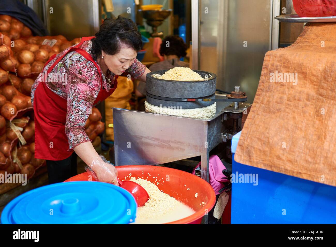 Eine Koreanerin in roter Schürze verwendet eine spezielle Mühle, um Mungbohnen für die Verwendung in Mungbohnenpfannkuchen (Bindae-tteok oder Bindaetteok) im Gwangjang MKT zuzubereiten. Stockfoto