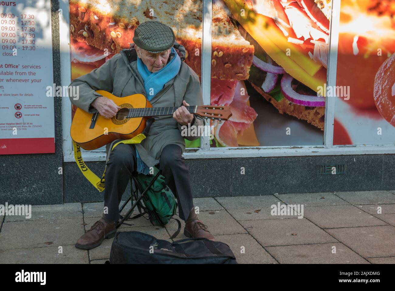 Alter Mann mit Gitarre ausserhalb des Co-op und Post auf Spring Gardens, Buxton, England Straßenmusik Stockfoto