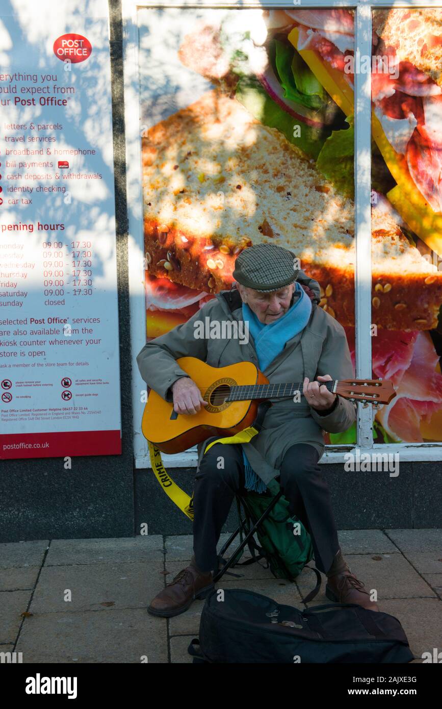 Alter Mann mit Gitarre ausserhalb des Co-op und Post auf Spring Gardens, Buxton, England Straßenmusik Stockfoto