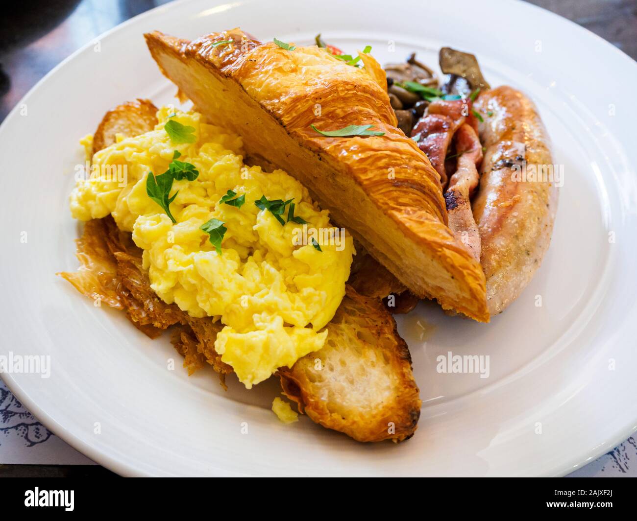 Ein ganztägiges Frühstück/Brunch combo Mahlzeit Teller mit Rührei, Croissants, Wurst, Speck und Pilzen auf eine weisse Platte bei einer Western casual Resta Stockfoto
