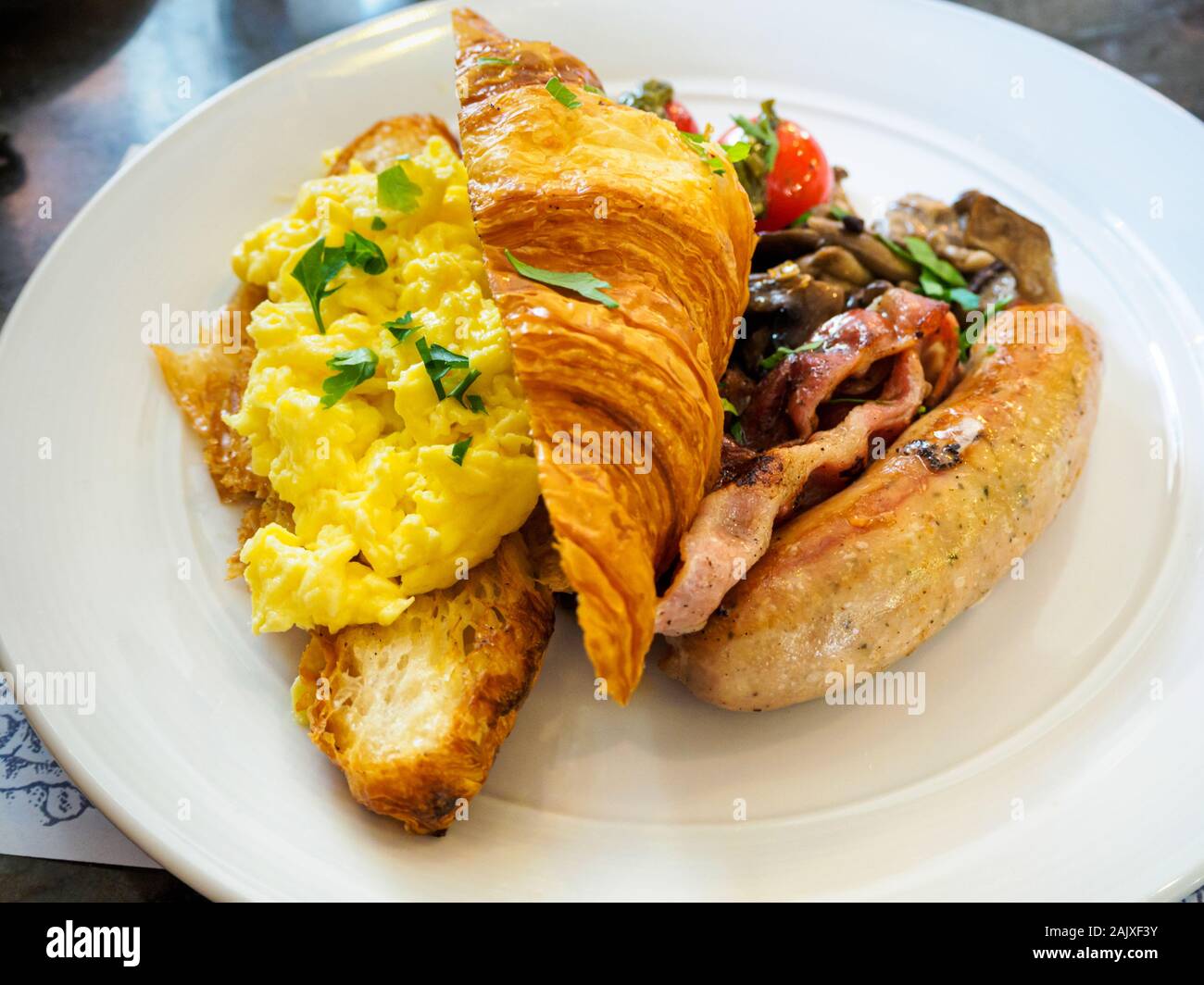 Ein ganztägiges Frühstück/Brunch combo Mahlzeit Teller mit Rührei, Croissants, Wurst, Speck und Pilzen auf eine weisse Platte bei einer Western casual Resta Stockfoto