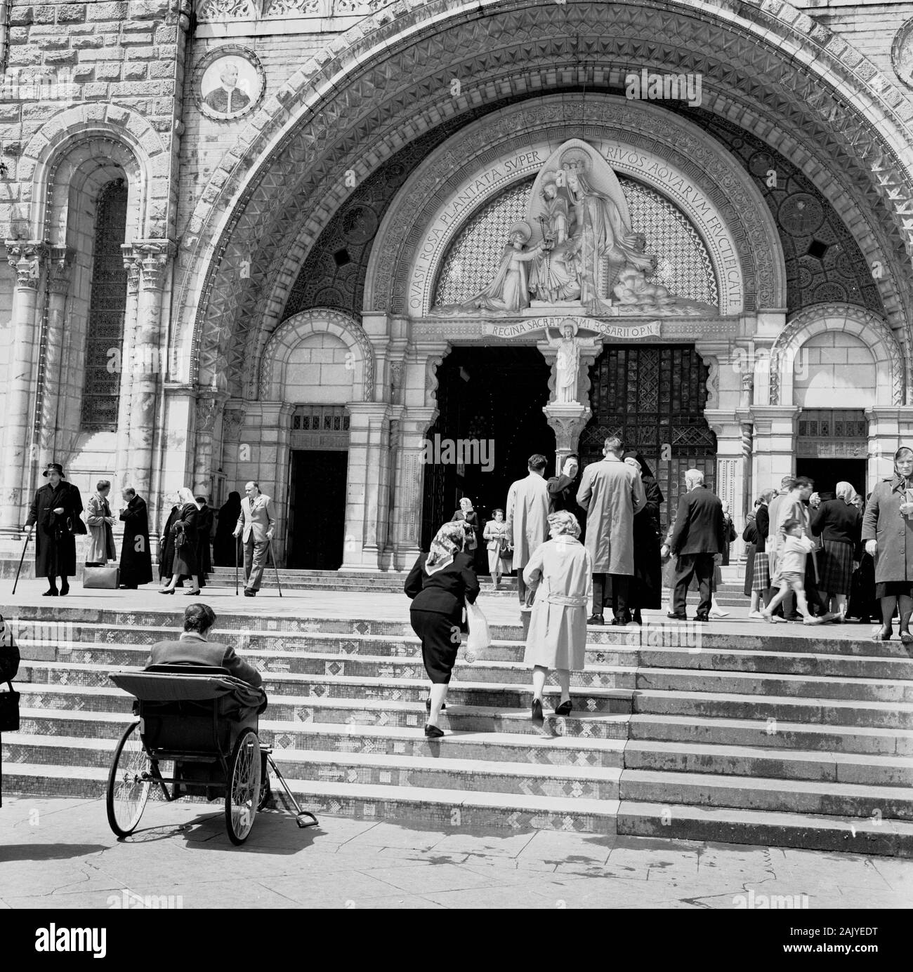 In den 1950er Jahren waren Besucher am Eingang zur Rosary Basilica, Lourdes, Frankreich, einer katholischen Kirche am "Heiligtum unserer Lieben Frau von Lourdes" oder der Domäne, einem Erdgebiet rund um den katholischen Schrein (Grotte), zu sehen. Ein Pilgerort für viele des christlichen Glaubens, vor allem für Kranke und Unfeste, wie ein Mann auf dem Bild in einer ungültigen Kutsche der Epoche an den Stufen der Kirche erkennen kann. Stockfoto