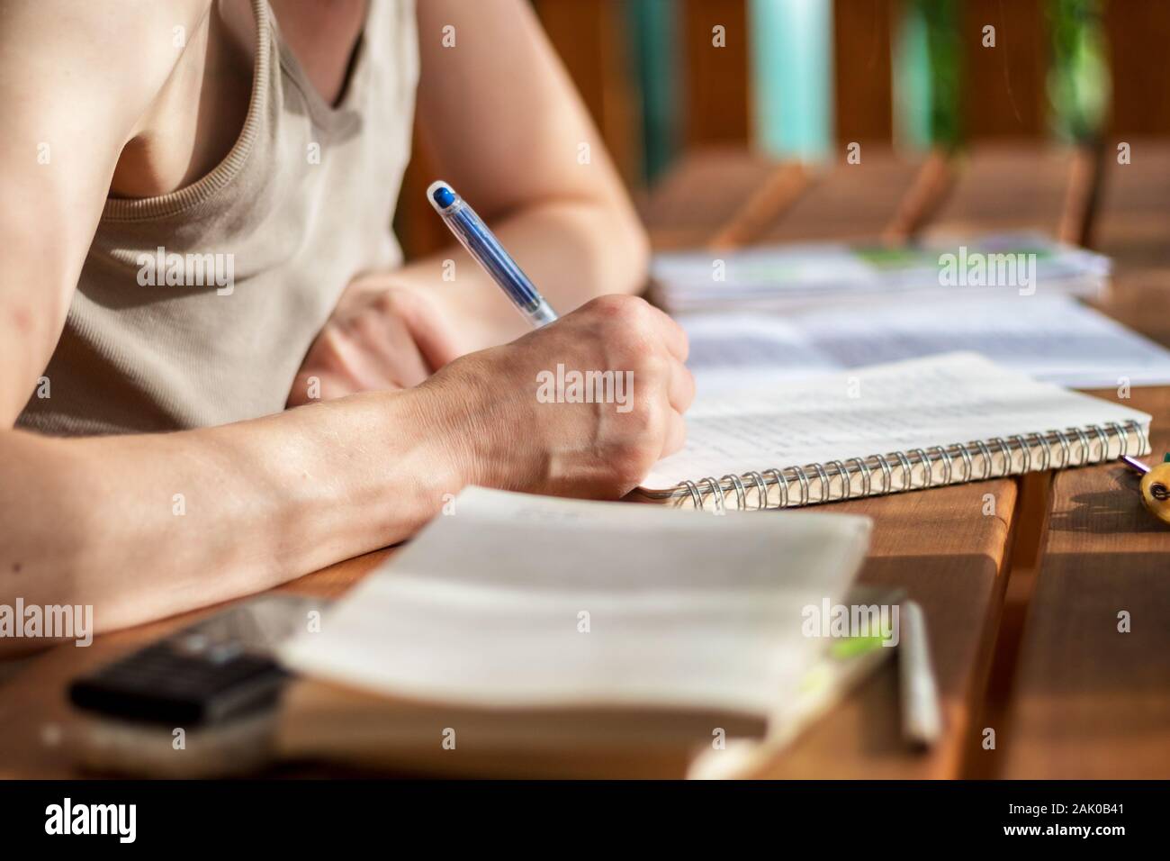 Nahaufnahme einer Frauenhand mit einem Stift, schreibt in ein Notizbuch, legt ein Buch auf den Tisch, Handy, Bleistifte, im Freien auf einer Terrasse auf einem sonnigen d Stockfoto