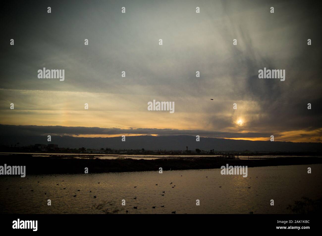Niedrige Sonne, die an einem bewölkten Wintertag, kurz vor Sonnenuntergang, direkt über den Bergen hinter einer riesigen Wasserfläche glüht. Sonne rechts vom Horizont. Stockfoto