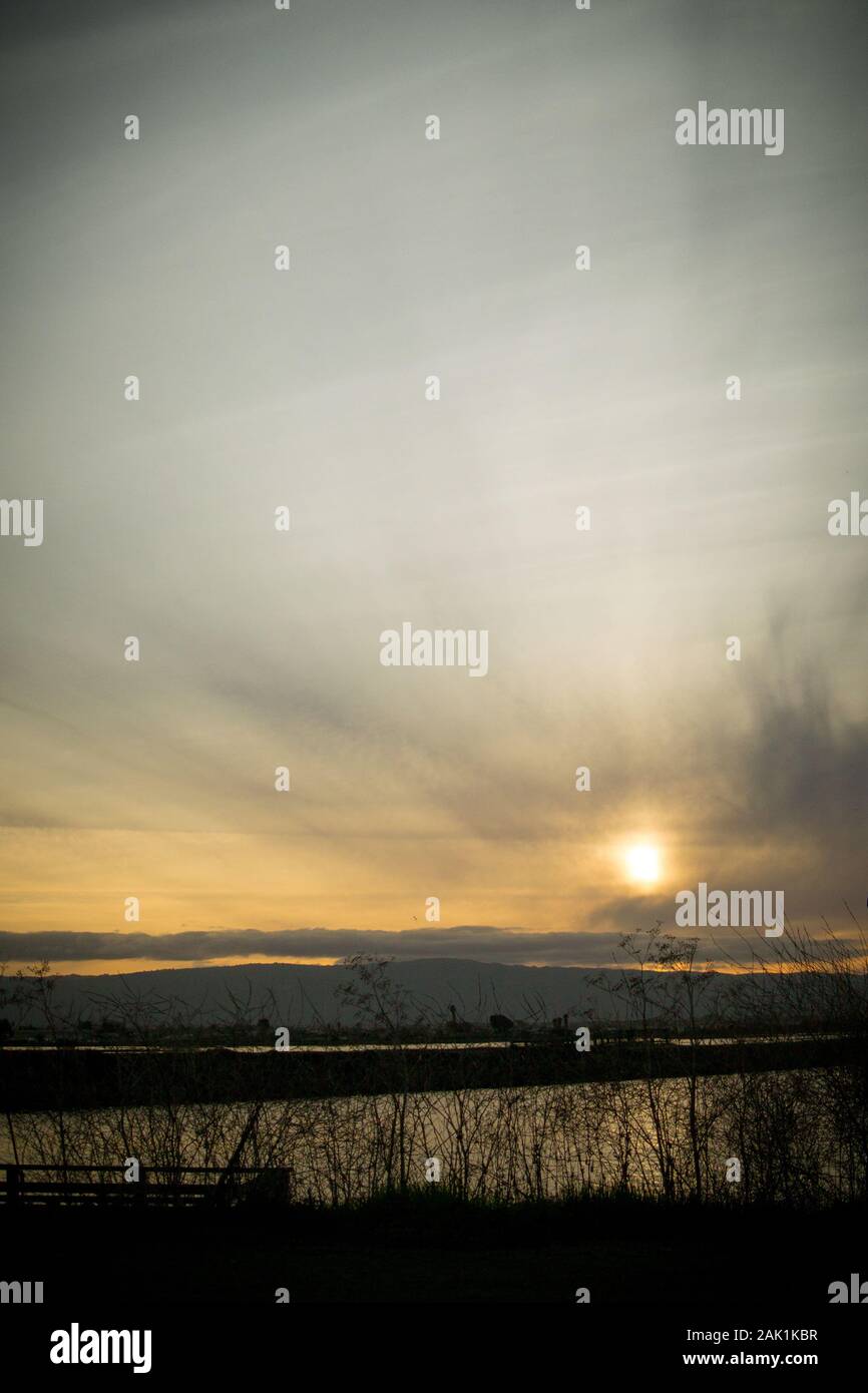Niedrige Sonne, die an einem bewölkten Wintertag, kurz vor Sonnenuntergang, direkt über den Bergen hinter einer riesigen Wasserfläche glüht. Stockfoto