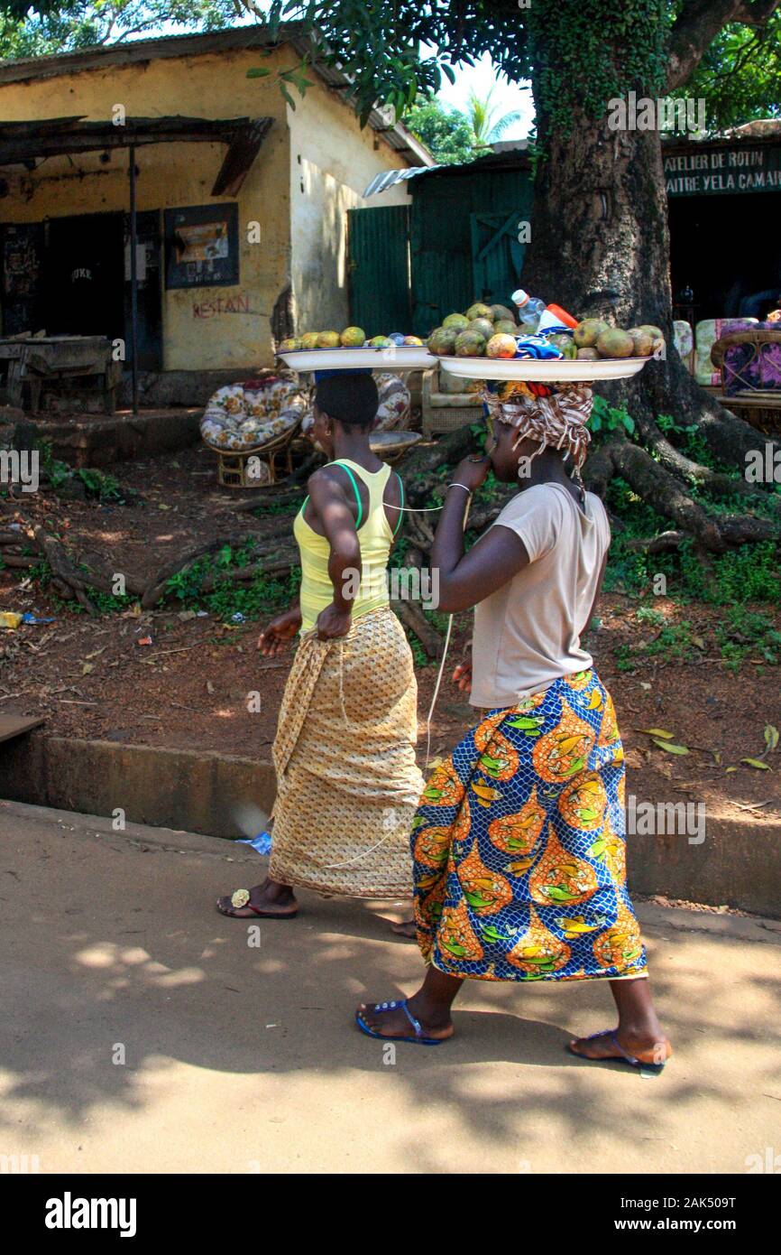 Afrikanische Frauen balancieren Körbe mit Früchten auf dem Kopf und gehen auf einer schmutzigen Straße in Conakry, Guinea, Westafrika Stockfoto