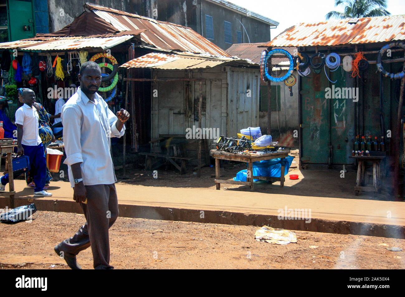 Afrikanischer Mann, der auf der Hauptstraße in Conakry, Guinea, Westafrika, vorbei an Marktständen und einer Tankstelle mit Treibstoff in Glasflaschen unterwegs ist Stockfoto