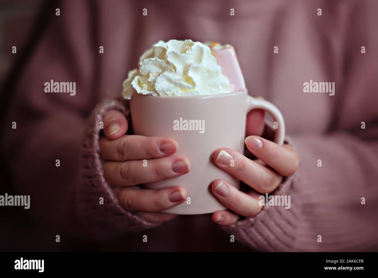 Rosa Tasse mit Kaffee mit Sahne und Marshmallows in die Hände einer Frau in einem warmen Rosa Strickpullover. Stockfoto