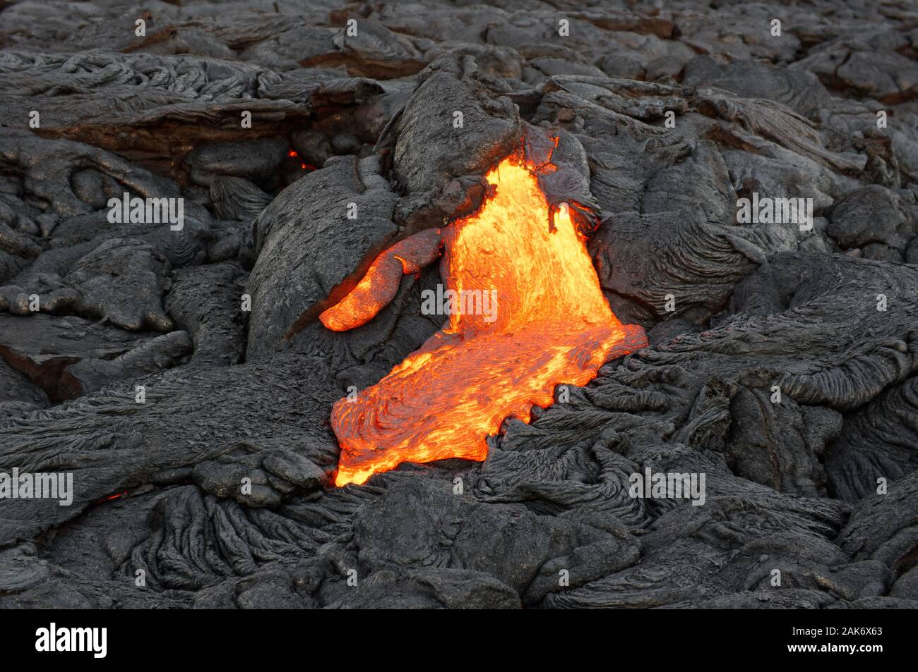 Heiße Magma eines Lavastroms aus einer aktiven vulkanischen Eruption taucht aus einer Spalte und fließt über zuvor hinterlegten dunkel stark strukturierte Felsen, Stockfoto