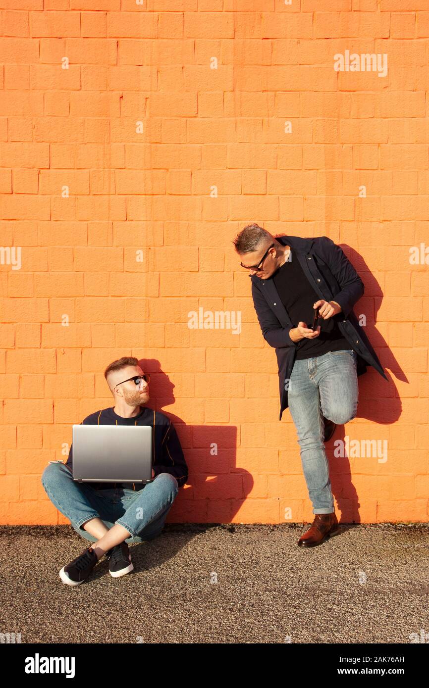 Zwei Freunde in casual-wear in städtischen Wettbewerb mit digitalen Geräten. Jungen kaukasischen Mann sitzt im Schneidersitz auf dem Boden und Arbeiten am Laptop. Die anderen Stockfoto