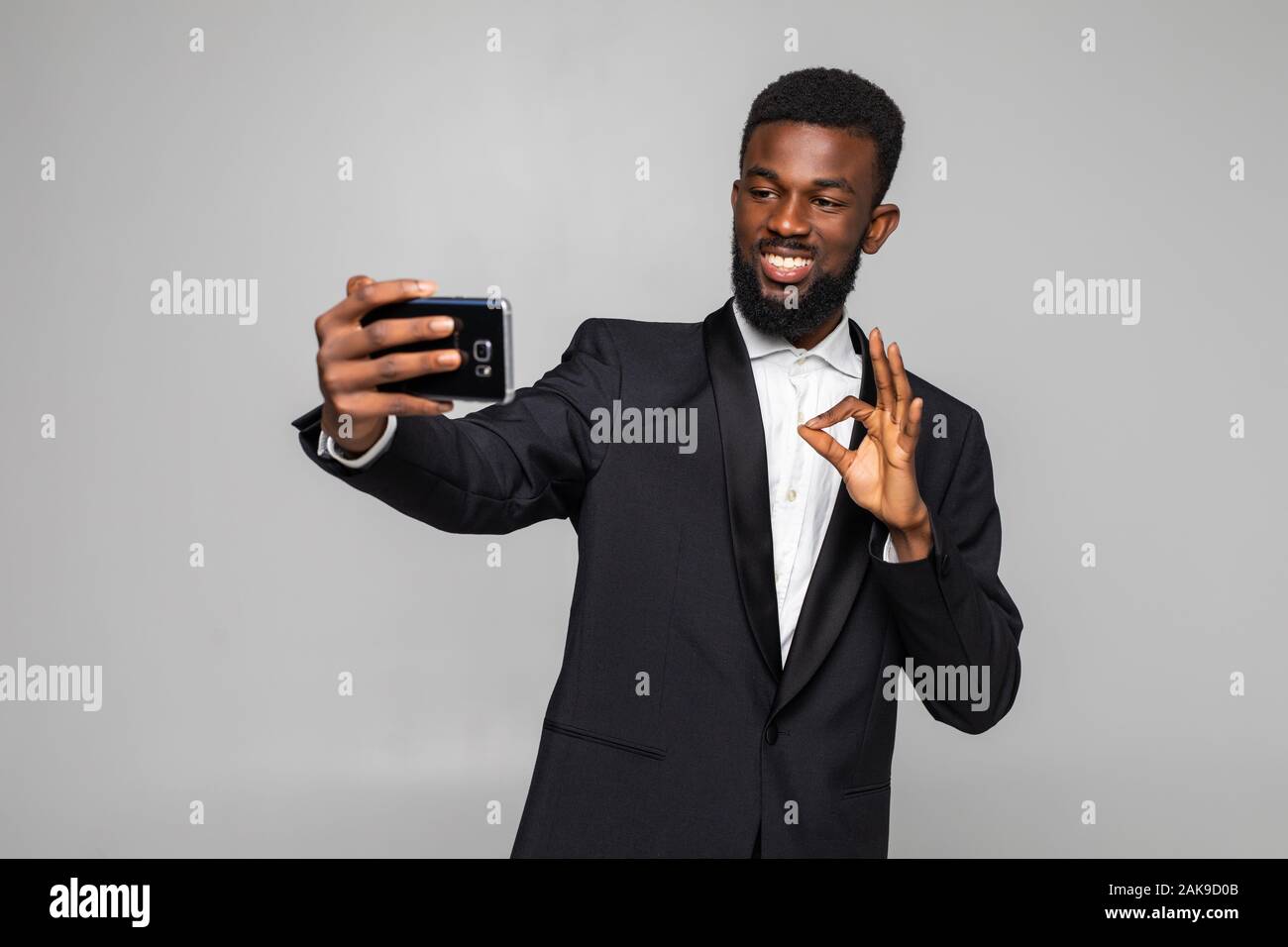 Junge afrikanische Mann in Anzug, selfie mit okay Geste auf grauem Hintergrund Stockfoto
