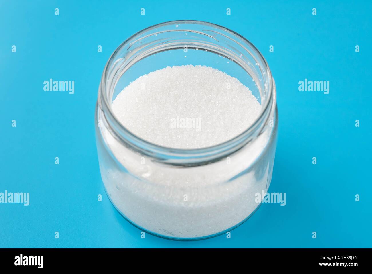 Ein Haufen Zucker in einem Glas auf blauem Hintergrund. Der runden transparenten Bank mit kristallinem Zucker Stockfoto