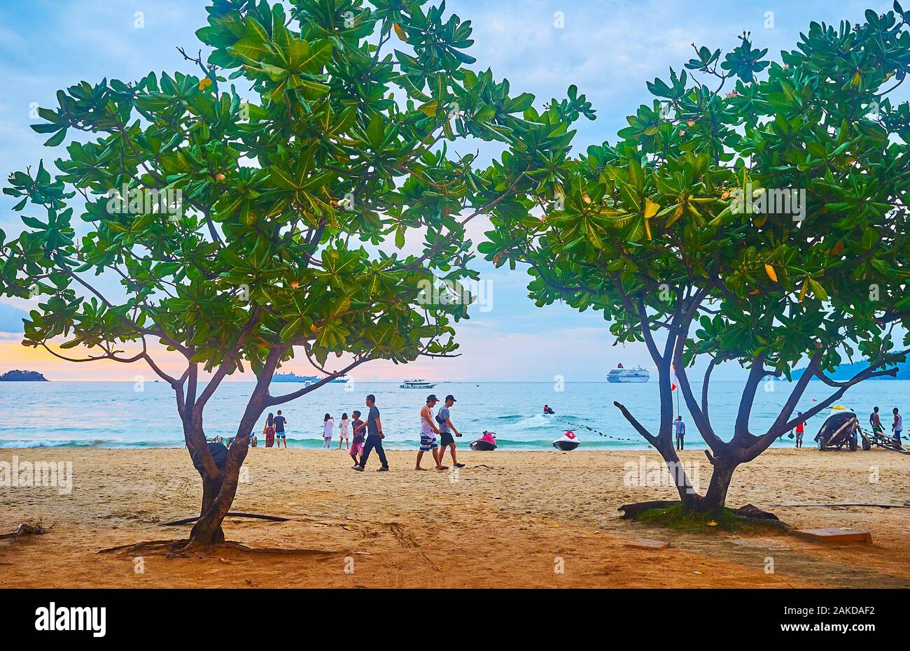 PATONG BEACH, THAILAND - 1. Mai 2019: Die Urlauber zu Fuß entlang der zentralen Strand des Resorts mit schattenspendenden Feigenbäumen und günstig Wasser Roller, am 1. Mai in P Stockfoto