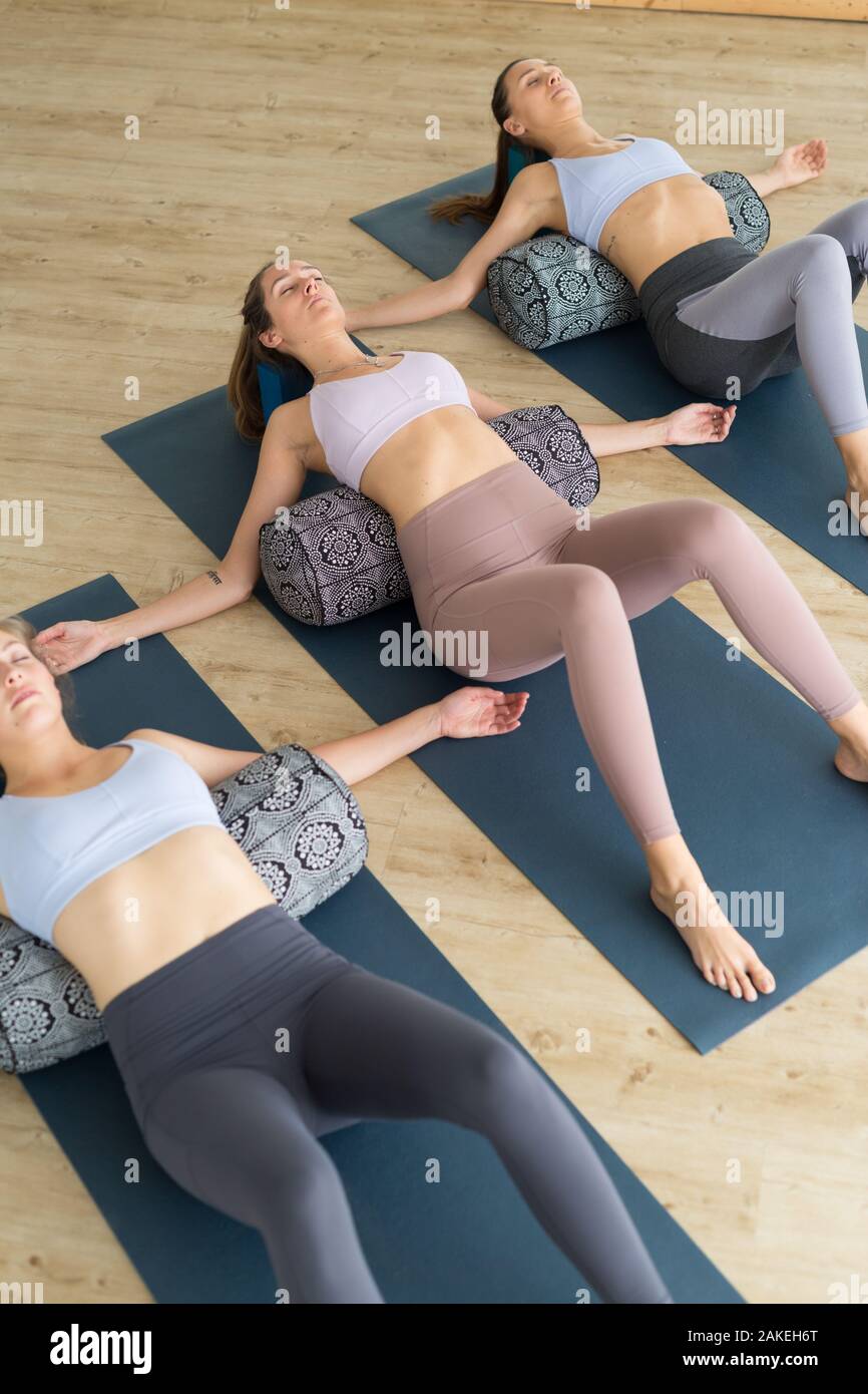 Restorative Yoga mit Polstern. Gruppe von drei Jungen sportlich attraktive Frauen in Yoga Studio, liegen auf Kissen Polstern, Stretching und Entspannung Stockfoto