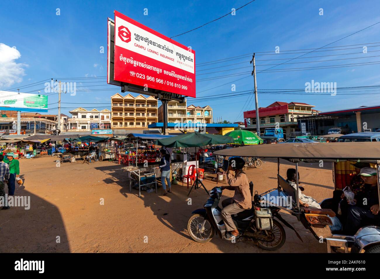 Ein tuk tuk Passagiere, die durch ein Verkehrsknotenpunkt mit Street Food gefüllt Stände Antriebe unter einer großen plakatwand in Skun, Kambodscha. Stockfoto