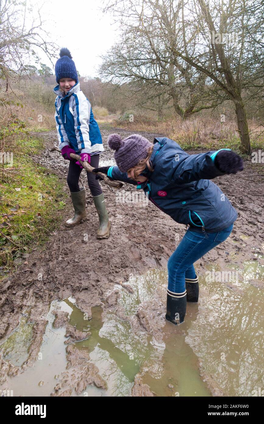 Zwei junge Mädchen/Mädchen/Schwestern im Alter von neun Jahren und 7 Jahren, die Wellington-Stiefel tragen, helfen gern, retten und spielen in einer großen nassen, schlammigen Pfütze während eines Familienuntergangs über West End Common, Esher, Surrey. GROSSBRITANNIEN (115) Stockfoto