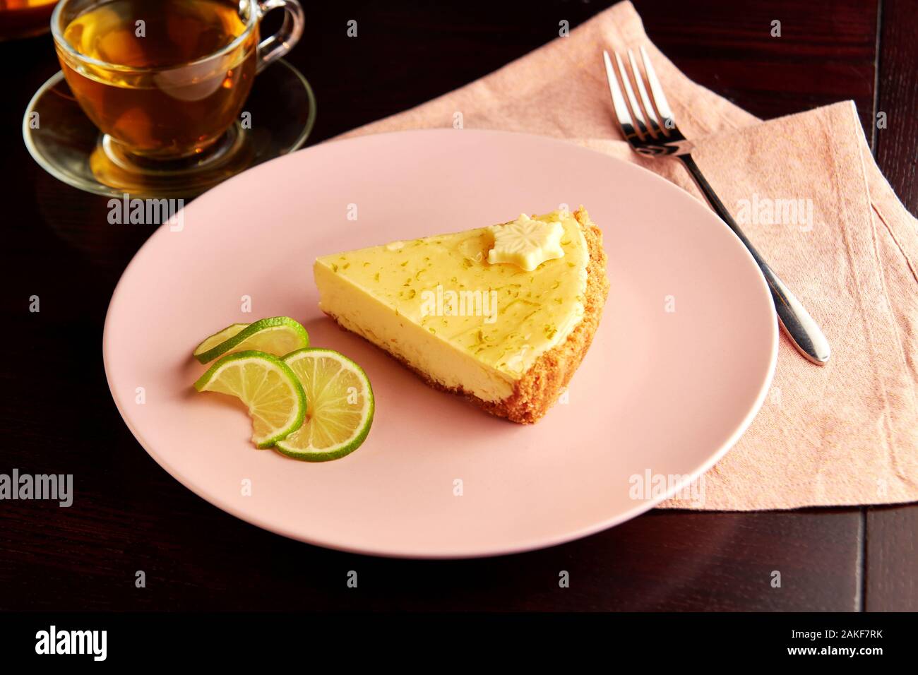 Stück leckeren Törtchen mit Limettenschale und Scheibe des Limes auf rosa Platte auf dunklen Holztisch, serviert mit Tasse Kräutertee. Stockfoto