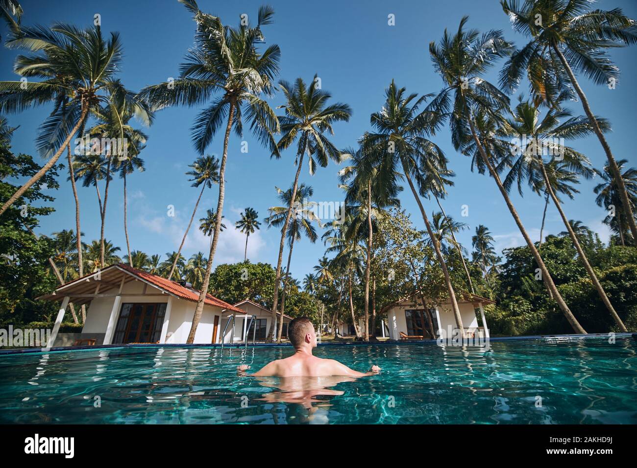 Entspannung im Pool inmitten von Kokospalmen. Junger Mann in Wasser, das gegen die Bungalows der Ferienanlage ruht. Stockfoto