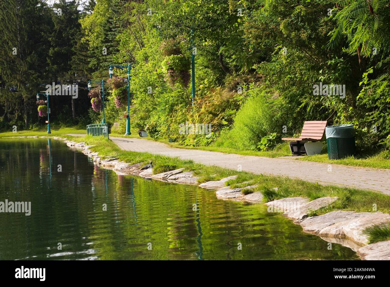 Stein umrandeten See mit Pfad und Strassenlaternen mit hängenden Körben von Violett Petunie im Sommer, Centre de la Nature öffentlichen Garten, Laval, Quebec, Kanada. Stockfoto
