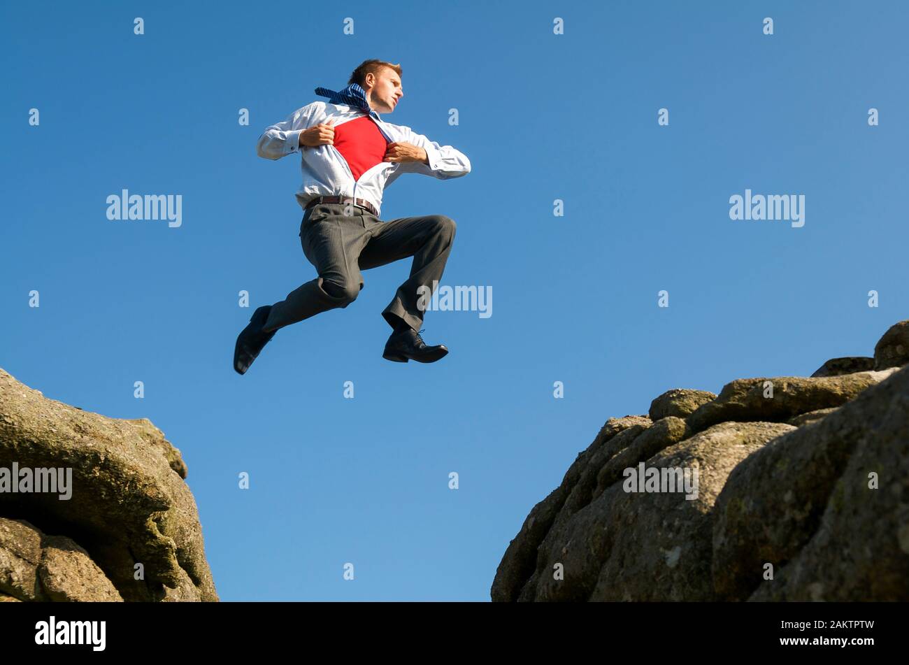 Superhelden Geschäftsmann mit einem gewagten Sprung zwischen zwei Felsbrocken im Freien gegen strahlend blauen Himmel Stockfoto