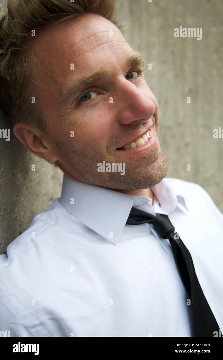 Porträt des Mannes in elegantem, legeren Outfit aus weißem Hemd und häutiger Rückenbinden lächelnd im Freien vor einer strukturierten Betonwand Stockfoto
