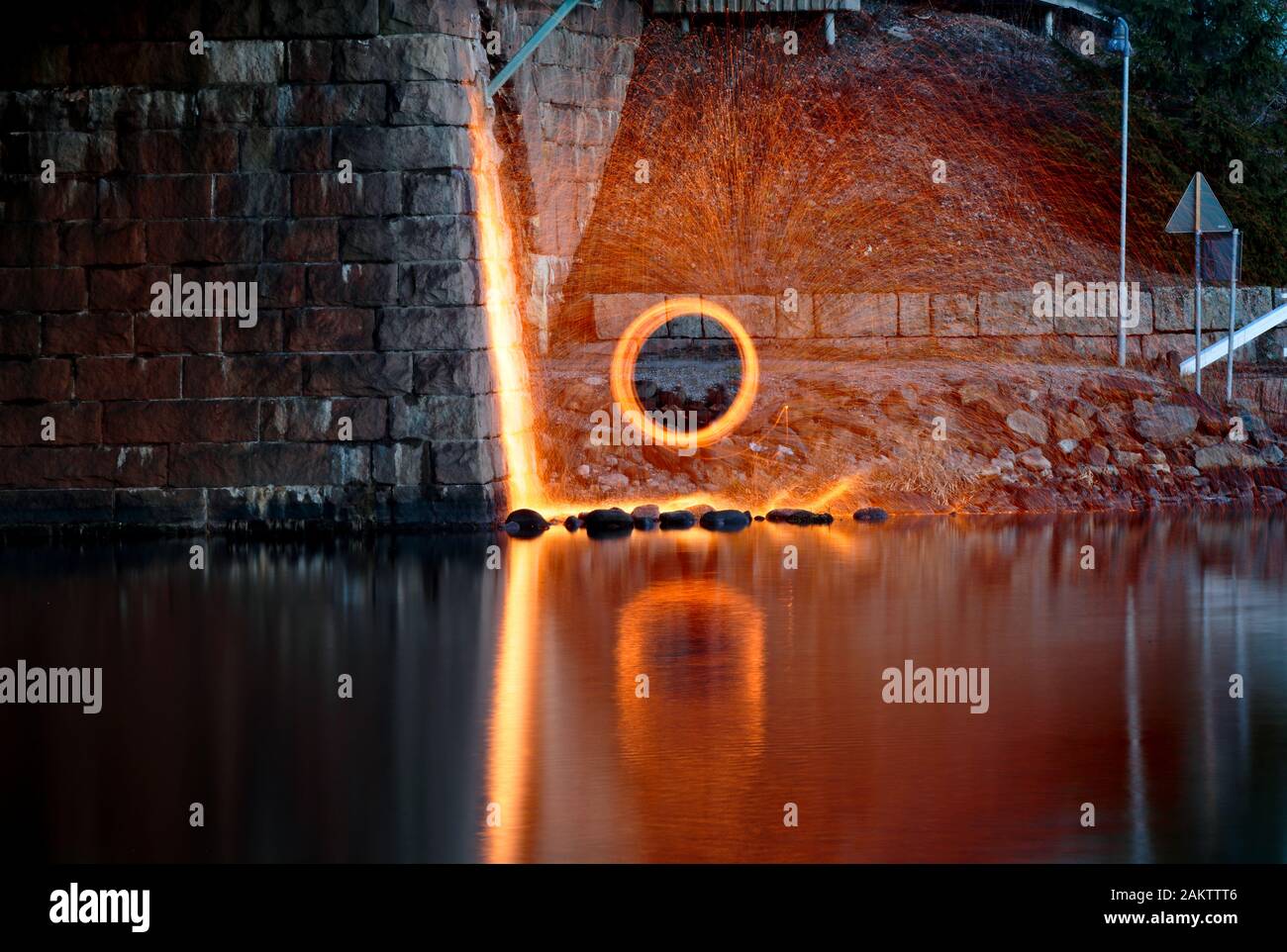 Feuerwerk aus brennender Stahlwolle, das sich neben einem See in der Nähe eines Brückenbauwerks dreht Stockfoto