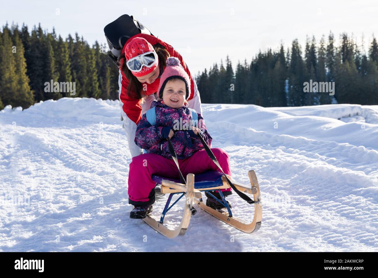 Adorable Girl Rodeln genießen, während ihre Mutter sie Schlitten gegen Pinienwald im Winter Stockfoto