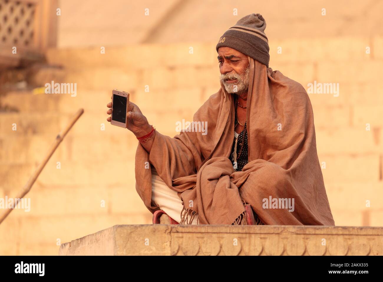 VARANASI, Indien, 18. Januar 2019: Ein Mann entlang einer Ghat sitzend und mit Blick auf den Ganges, hält ein Smartphone. Stockfoto