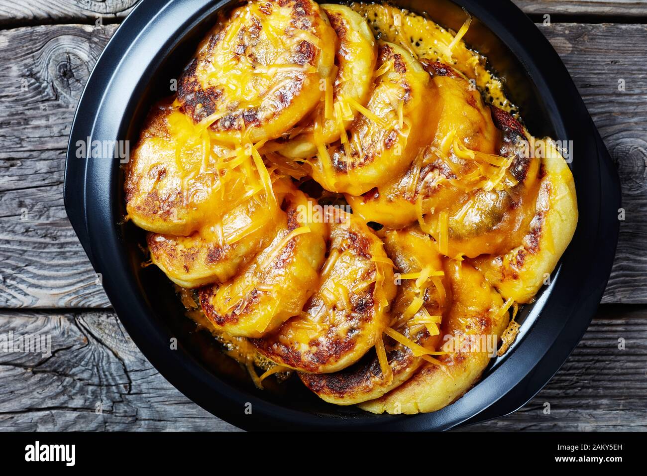 Hausgemachte Kartoffelkuchen mit geriebenem Cheddar-Käse in einem schwarzen Gericht auf einem rustikalen Holztisch, flach und nah Stockfoto