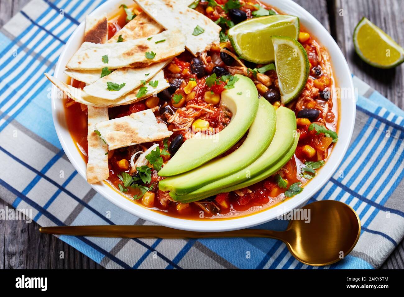 Chicken Taco Suppe mit schwarzer Bohne, Maiskörner, mit Tortillastreifen, Avocado-Scheiben und Kalk in weißer Schüssel auf einem rustikalen Holztisch mit Gold bestockt Stockfoto