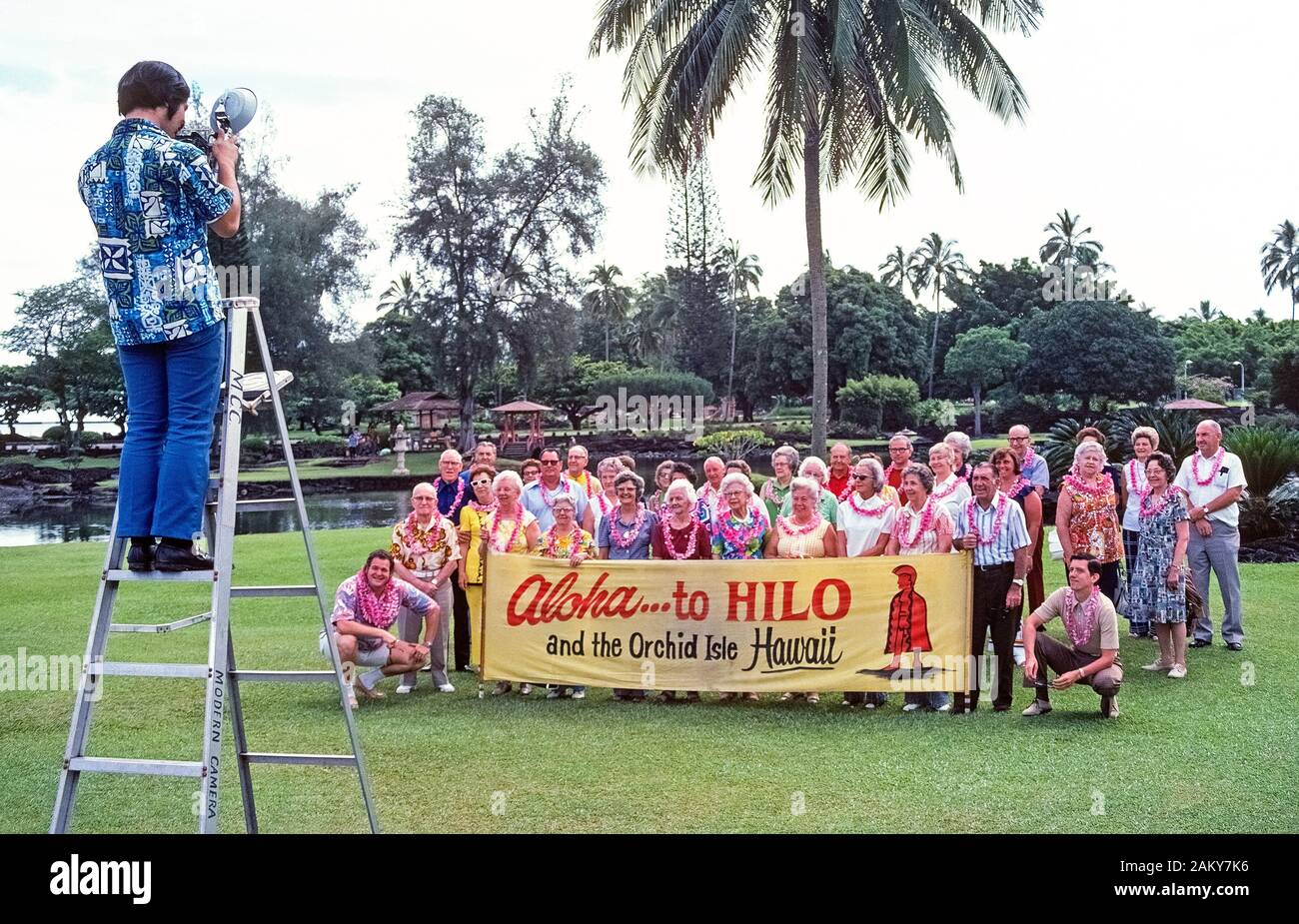 Eine Tour Gruppe der älteren Amerikaner sammelt hinter ein Zeichen für ein offizielles Foto in Hilo auf Big Island am Anfang von einem Besuch in den Pazifischen Ozean Inseln von Hawaii, USA zu stellen. Sowohl Männer als auch Frauen tragen frische Blumenkränze durch Hosts der Tour unternehmen als die traditionellen Hawaiianischen 'Aloha' Willkommen zu den Besuchern präsentiert. Diese Urlauber waren im Jahr 1974 fotografierte an einem bewölkten Tag im liliuokalani Park, dass Grenzen Hilo Bay und verfügt über japanische Gärten. Historisches Foto. Stockfoto