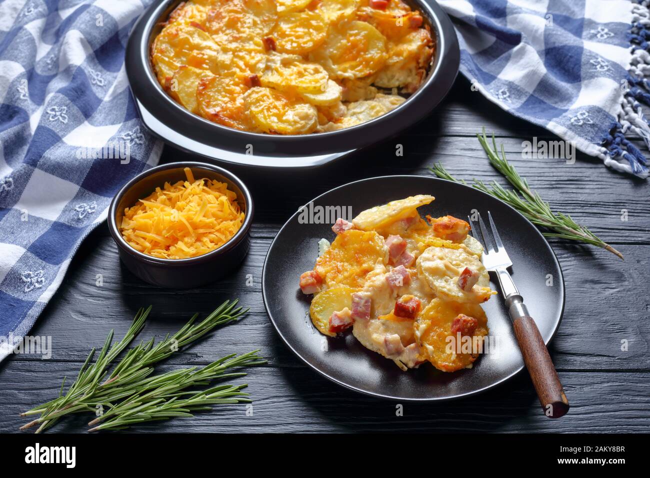 Nahaufnahme von geschälten Kartoffeln, mit Cheddar-Käse und Ham in einer Backform und auf schwarzem Teller serviert, Ansicht von oben, Stockfoto