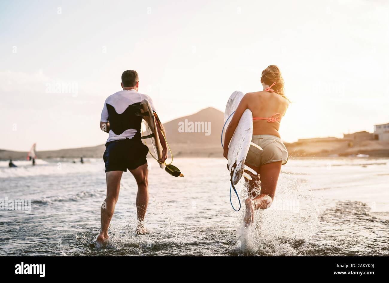 Fröhliche Surfer, die bei Sonnenuntergang im Wasser laufen - Junge Paare haben Spaß beim Surfen im Meer - Lifestyle-Konzept für Extremsport und Jugendkultur Stockfoto
