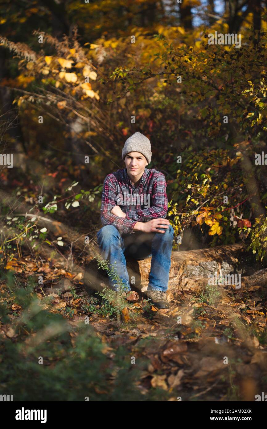 Selbstbewusster Teenager-Junge in karierten Hemd und Jeans sitzt auf Log in Holz. Stockfoto
