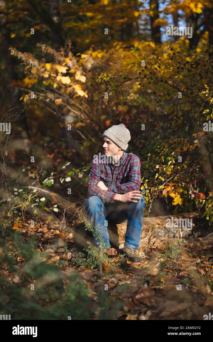 Teenager Junge sitzt allein auf Log in Wald und lächelt mit gekreuzten Armen Stockfoto