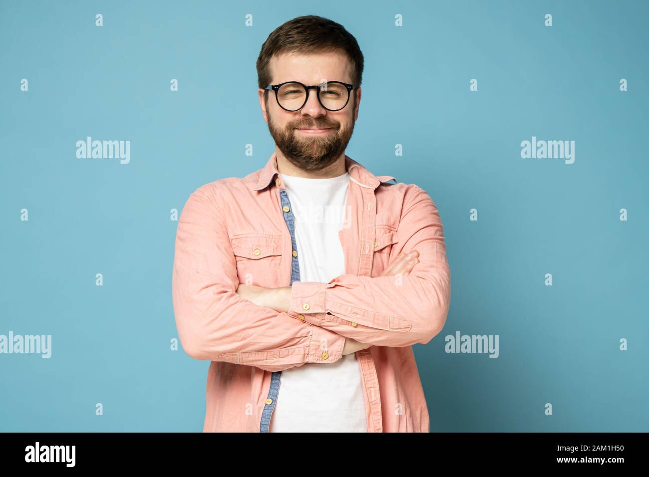 Lustiger bärtiger Mann mit Brille steht mit gekreuzten Armen, lächelt seltsam und blickt mit verengten Augen auf die Kamera. Stockfoto