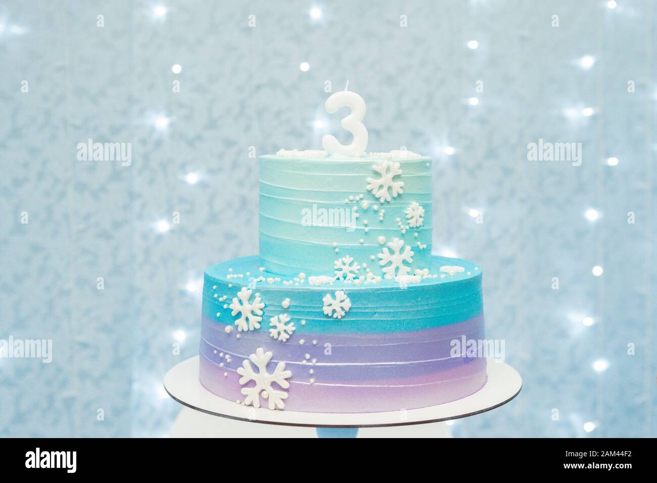 Kuchen zeigen Details von wunderbarem blauem und violettem Geburtstagskuchen, der mit Schneeflocken dekoriert ist. Zweistufiger Kuchen auf blauem Hintergrund mit unfokussierten Lichtern. Stockfoto