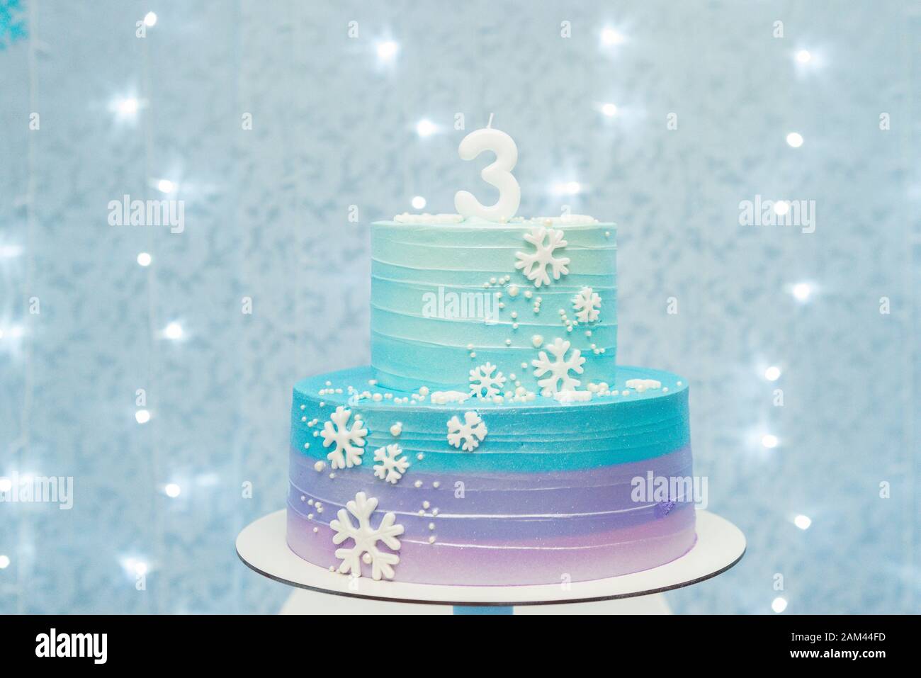Kuchen zeigen Details von wunderbarem blauem und violettem Geburtstagskuchen, der mit Schneeflocken dekoriert ist. Zweistufiger Kuchen auf blauem Hintergrund mit unfokussierten Lichtern. Stockfoto