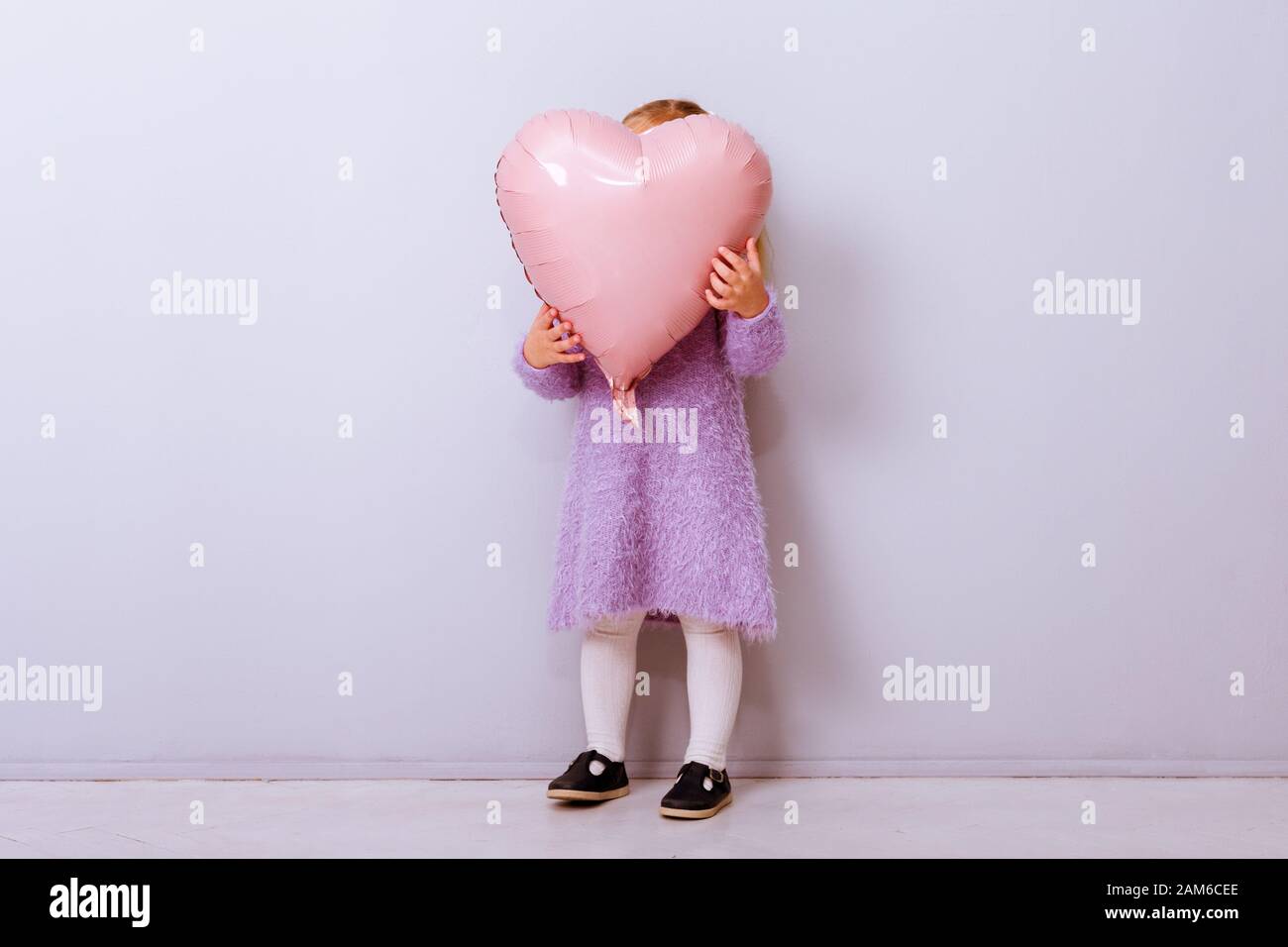 Glückliches Herz des Valentinstag. Baby Mädchen bedeckte ihr Gesicht mit einem rosafarbenen Herzballon auf einem hellvioletten Hintergrund. Stockfoto