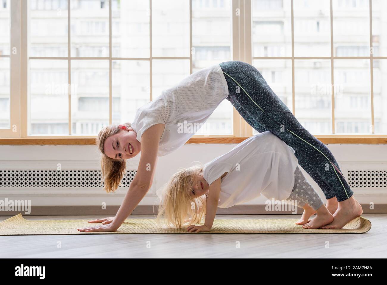 Junge Mutter macht Yoga mit 3-jähriger Tochter vor dem Fenster. Glücklich Mama lächelt während Yoga zusammen mit ihrem niedlichen Mädchen zu üben Stockfoto